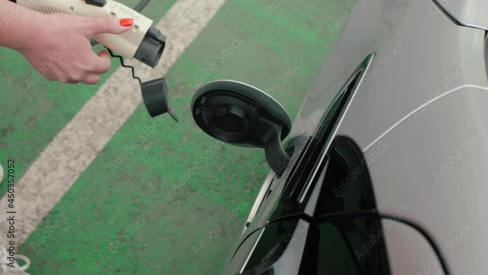 Female hands unplug electric car power supply cable to charge EV and close charging port lid, close up on green parking lot. Unrecognizable woman in red dress with white dots disconnects plug Female hands unplug electric car power supply cable to charge EV and close charging port lid, close up on green parking lot. Unrecognizable woman in red dress with white dots disconnects plug