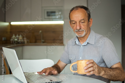 Trendy mature man is working from home with laptop sitting at the table in his kitchen, happy retirement, home-office concepts