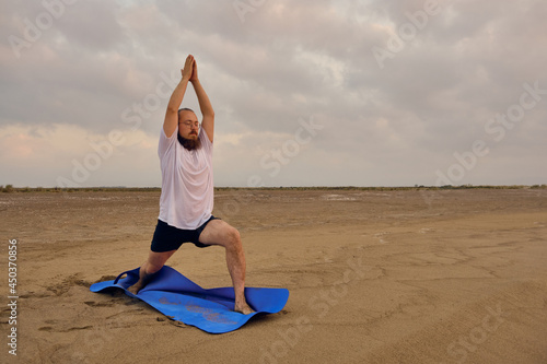 Man stands on blue exercise man doing virabhadrasana one pose. Man practices warrior one asana at sea beach during sunrise photo