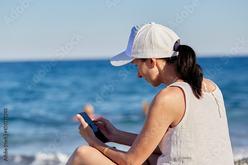  Young woman in a hat with a phone on the beach near the sea. The concept of freelancing, working online, online conference or studying online.