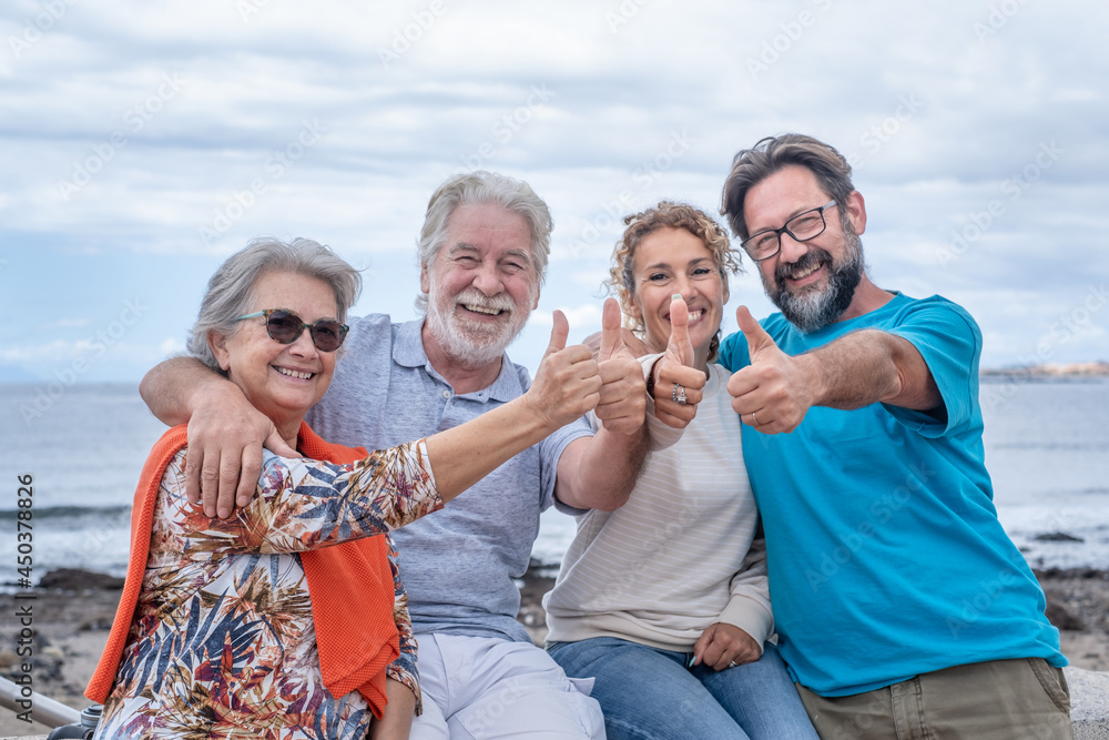 Multi generation family sitting together at the sea looking at camera with positive gesture, two happy and smiling couples