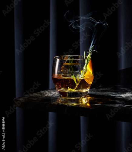 Smoked Whiskey Cocktail with Smoking Lavender Rosemary on Black Granite Bar Table Top with Dark Curtain Background.  Orange peel garnish and large square ice cube.