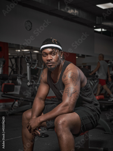 African american male fitness instructor sitting on bench. Handsome powerful athletic man in stylish sportswear in gym © Georgii