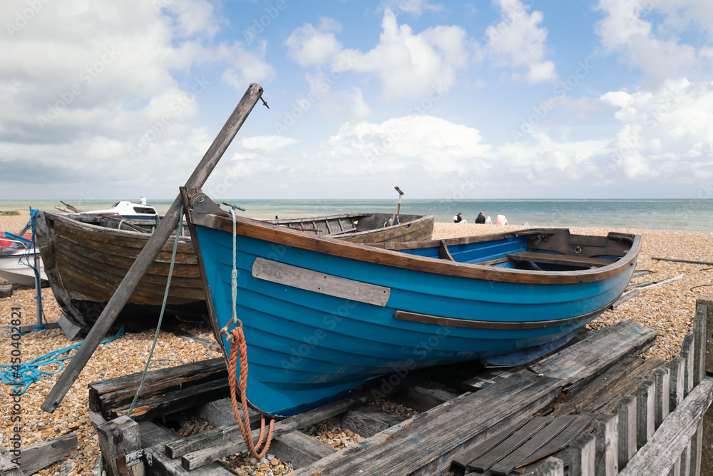 A quaint small blue wooden rowing boat berthed on wooden beams on a pebble beach next to another wood boat. A few people can be seen sitting on the beach by the sea behind the boat.