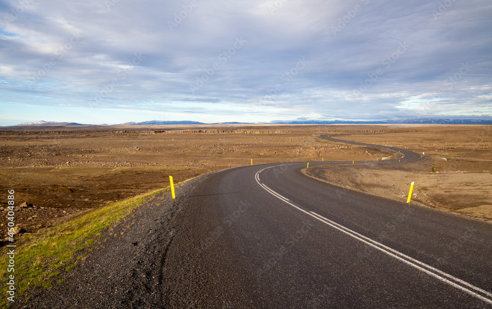 Asphalt road zigzags to the horizon among yellow fields under blue sky. Iceland.