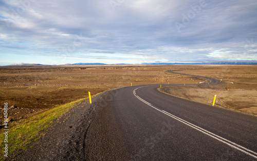 Asphalt road zigzags to the horizon among yellow fields under blue sky. Iceland.