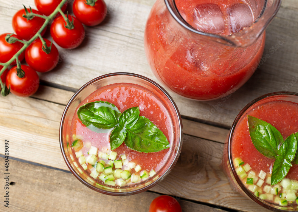Top view of gazpacho soup in glass glasses and a jug of ice on a wooden table. Cherry tomato branch, basil leaves and fresh cucumber, sliced brunois Top view of gazpacho soup in glass glasses and a jug of ice on a wooden table. Cherry tomato branch, basil leaves and fresh cucumber, sliced brunois