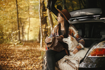 Stylish woman traveller with backpack sitting with cute dog in car trunk in sunny autumn woods. Young hipster female traveling with swiss shepherd white dog. Travel and road trip with pet