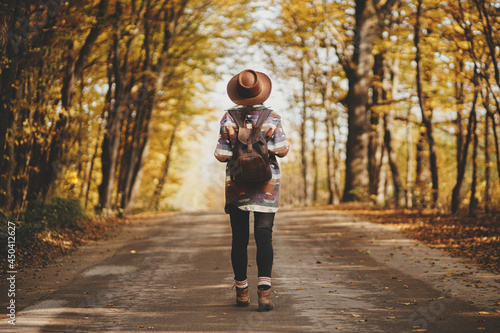 Stylish woman traveller in hat with backpack walking on road in sunny autumn woods. Young female hipster hiking in fall forest, beautiful moment. Travel and wanderlust concept. Back view © sonyachny