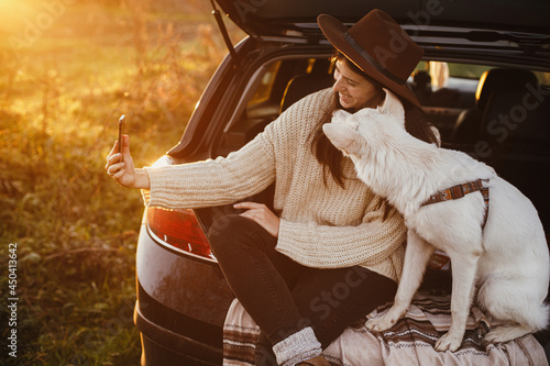 Autumn road trip with pet. Stylish hipster woman taking selfie photo with cute dog in car trunk in warm sunset light. Young female in hat and sweater using phone and travelling with sweet white dog