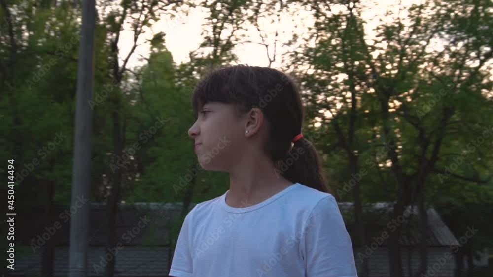 The teenage girl turns her head. Dressed in a white T-shirt. Evening time. Green trees in the background. Playground. City block. Leisure park. Slow motion. The teenage girl turns her head. Dressed in a white T-shirt. Evening time. Green trees in the background. Playground. City block. Leisure park. Slow motion.