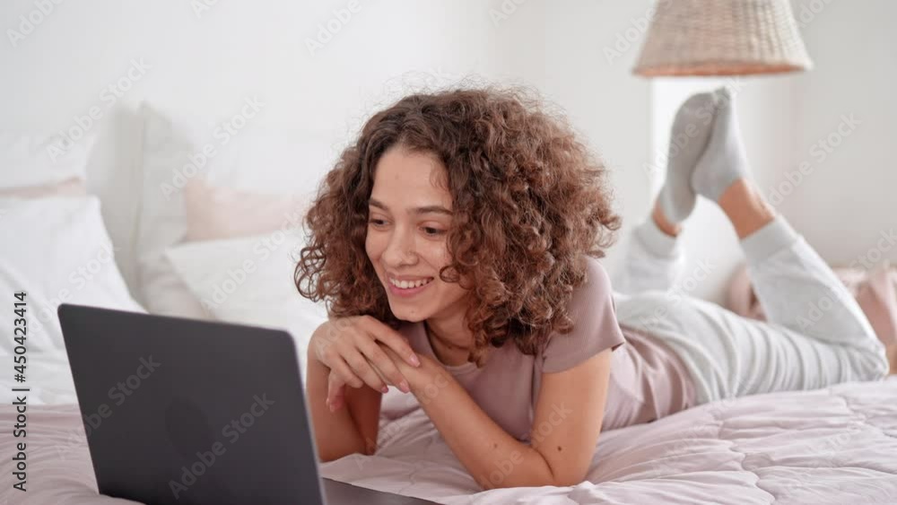 brunette with curly hair lying on bed has video call waving hand cheerful smiling communication with webcam app for calling home interior brunette with curly hair lying on bed has video call waving hand cheerful smiling communication with webcam app for calling home interior