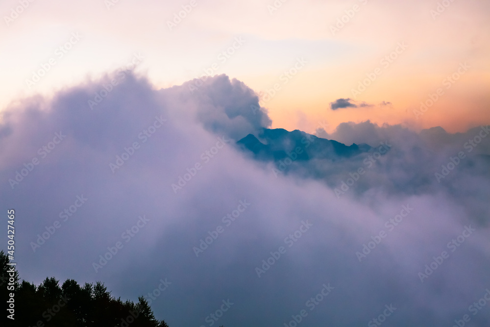 Dark Caucasus mountains peak shrouded in fog and fluffy clouds at colorful sunset. Pink and yellow cloudy skyscape. Stunning tranquil scene in Europe. Beauty in peaceful nature.
