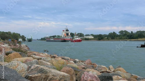 Large gray cargo ship entering the Port of Liepaja in calm sunny summer day, stone pier in foreground, Oskara Kalpaka swing bridge in background, waves splashing, wide shot photo