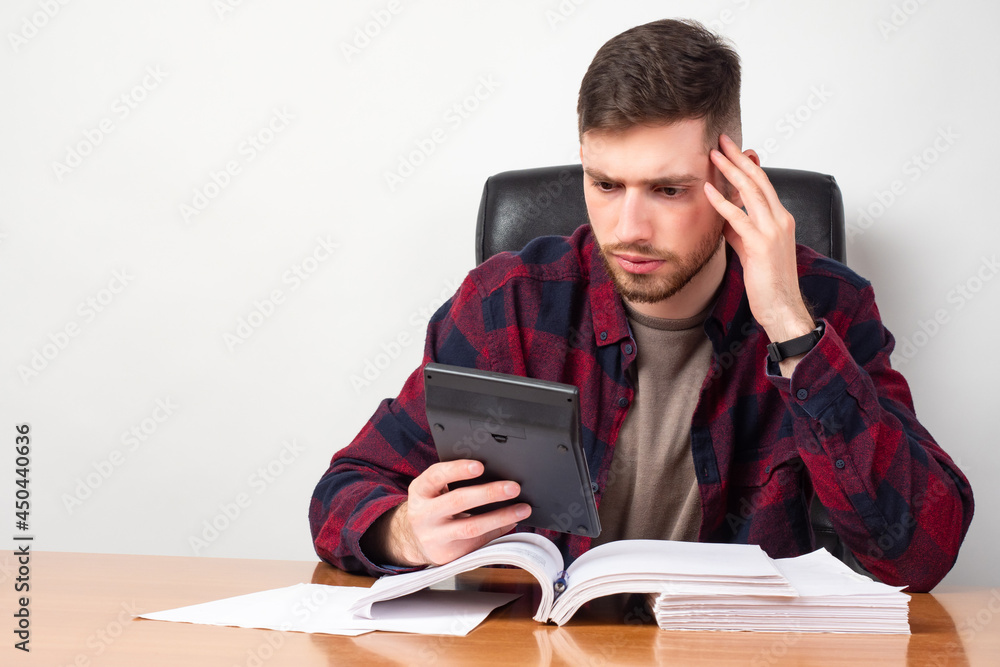 Man in amazement looks at the calculator on a light background. The calculation revealed losses. Financial audit shows losses. Man in amazement looks at the calculator on a light background. The calculation revealed losses. Financial audit shows losses.