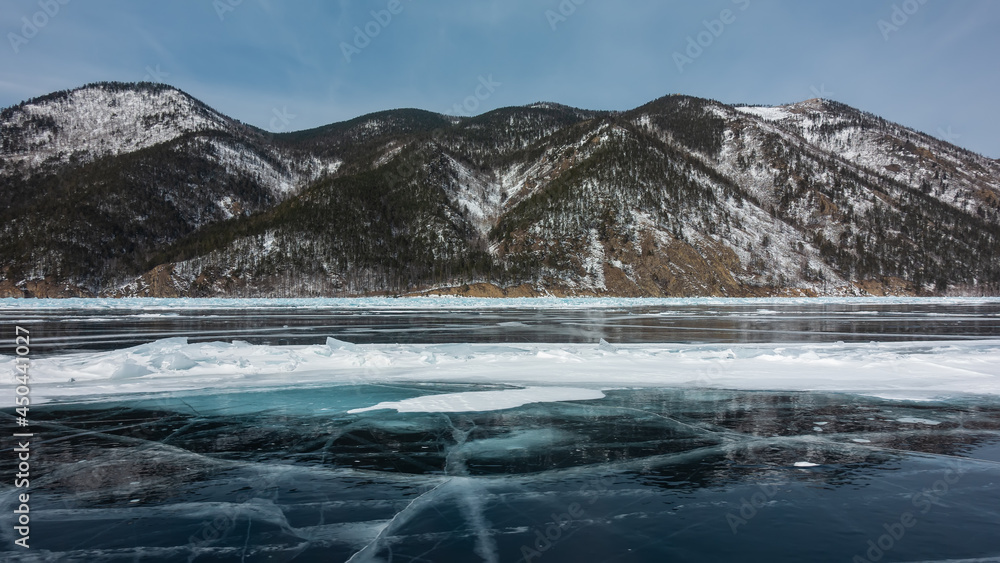 Cracks, snow spots, hummocks are visible on the blue ice of the frozen lake. The wooded slopes of the coastal mountains are covered with snow . Reflection on a smooth surface. Winter day. Baikal