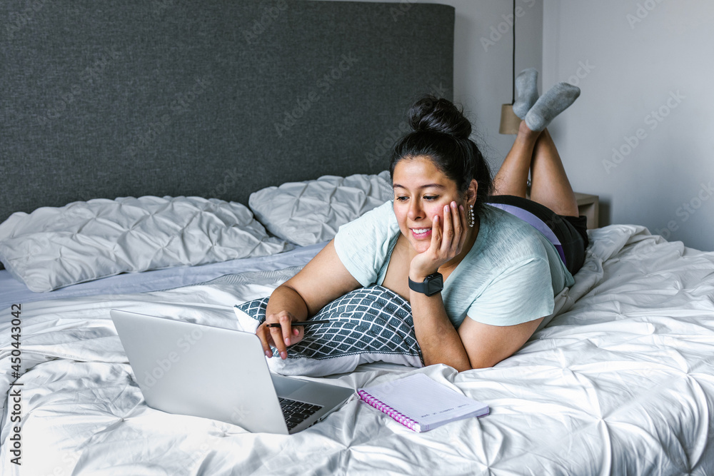 Fototapeta premium Curvy latin woman lying on bed using computer in Latin America, plus size female