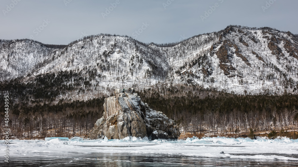 A granite rock with bizarre outlines and a flat top rises above a frozen lake. There is snow at the base, hummocks. Reflection on the ice. The background is a snow-covered mountain range. Baikal