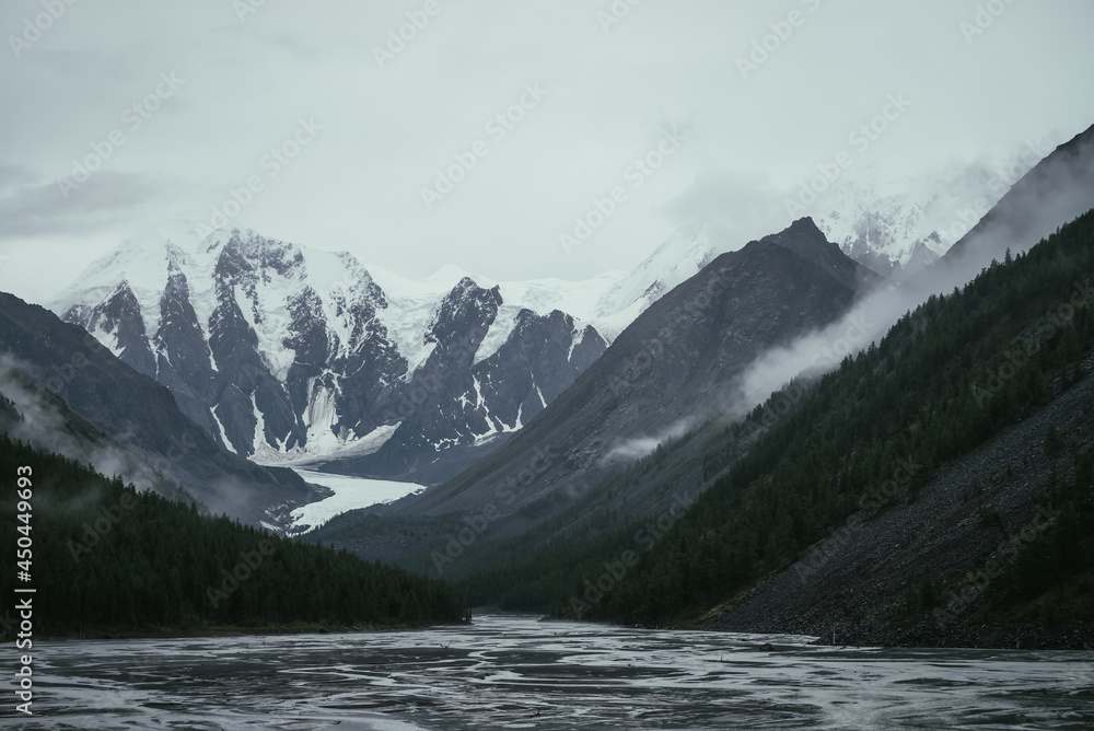 Atmospheric alpine landscape with mountain streams from snowy mountains in overcast weather. Bleak monochrome scenery with glacier tongue in mountain valley in rainy weather. Glacier among low clouds.