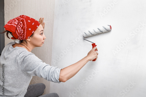 Asian woman applying primer and glue with a wide roller on the plastered surface of the wall for wallpapering