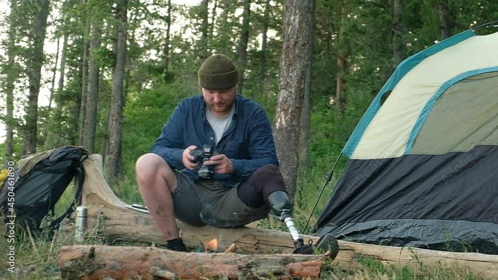 Young male tourist with prosthetic limb is resting by campfire in forest after long walk with backpack. disabled person is sitting on log using camera to create beautiful photos. Outdoor activities Young male tourist with prosthetic limb is resting by campfire in forest after long walk with backpack. disabled person is sitting on log using camera to create beautiful photos. Outdoor activities