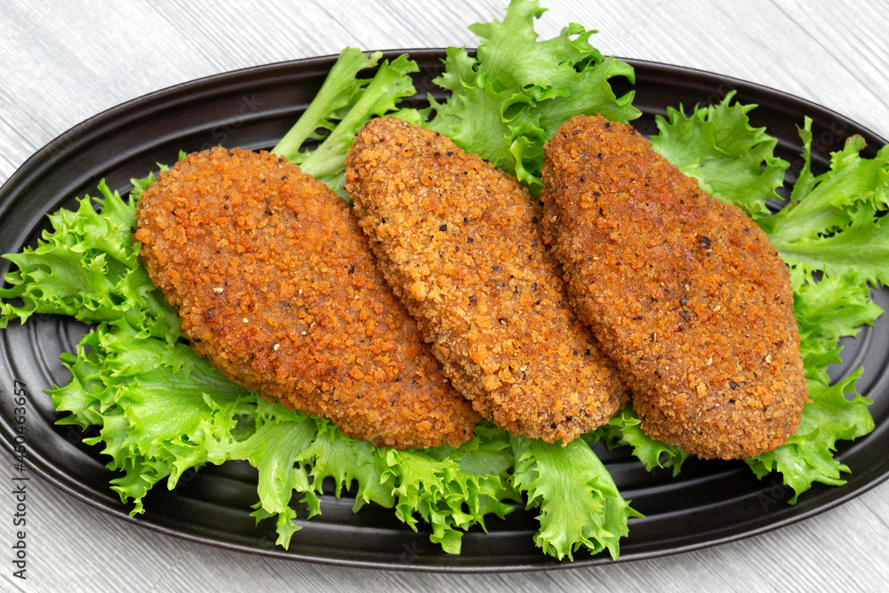 Southern fried chicken breast with spicy breadcrumbs on a bed of Lollo Biondo lettuce. On a black plate with a grey wood background Southern fried chicken breast with spicy breadcrumbs on a bed of Lollo Biondo lettuce. On a black plate with a grey wood background