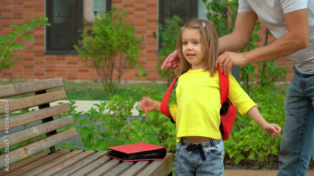 Father helps little daughter to put on school backpack in schoolyard. Happy schoolchild goes to school with textbooks. Education and preschool concept. Father helps little daughter to put on school backpack in schoolyard. Happy schoolchild goes to school with textbooks. Education and preschool concept.