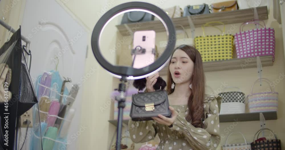 Slow motion of online seller Asian woman with long brown hair is broadcasting live on mobile phone with ring light, selling women's bags in her room against the backdrop of women's bag racks. Slow motion of online seller Asian woman with long brown hair is broadcasting live on mobile phone with ring light, selling women's bags in her room against the backdrop of women's bag racks.