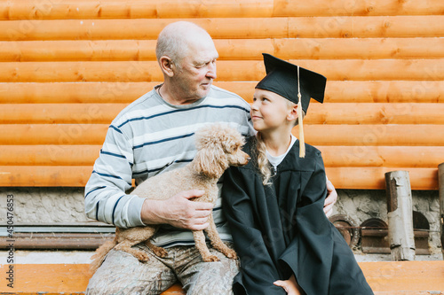 senior elderly man grandfather holds poodle dog in his arms and hugs his grandson boy who graduate of elementary school in cape and graduate cap on porch of rustic wooden house, concept of graduates