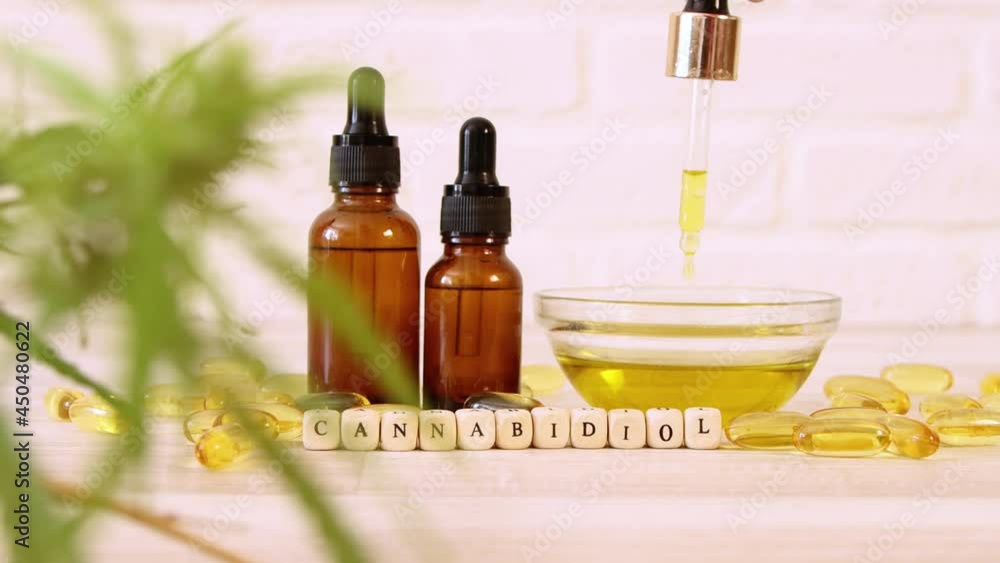 Hand dropping CBD oil from pipette to the bowl surrounded by jars, capsules and Cannabis plant. Concept of herbal alternative medicine, cbd oil, pharmaceutical industry. Hand dropping CBD oil from pipette to the bowl surrounded by jars, capsules and Cannabis plant. Concept of herbal alternative medicine, cbd oil, pharmaceutical industry.