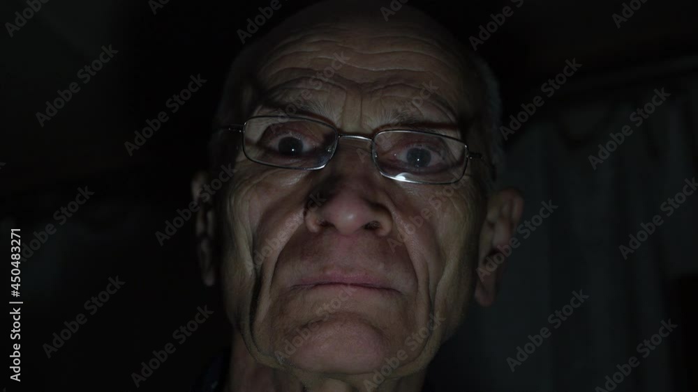 Senior man with wrinkles and glasses looks straight with angry face under bright electric light in darkness at home close view Senior man with wrinkles and glasses looks straight with angry face under bright electric light in darkness at home close view