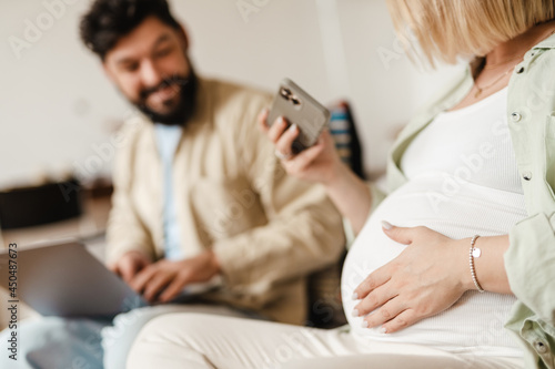 Man and pregnant woman using cellphone and laptop while sitting on couch