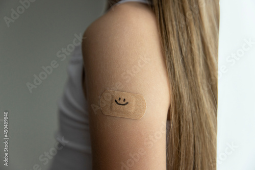 Little girl with long brown hair in white tank top got her vaccine, little's girl arm with plaster with drawn smile after vaccine