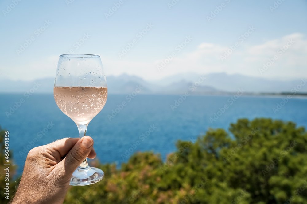 Glass of wine in hand. A glass of rose wine against the background of the Mediterranean sea and blue sky on a sunny summer day. Summer, travel, lifestyle, relaxation, and enjoyment concept.