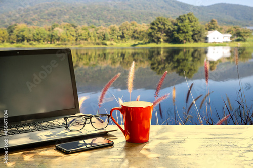Red coffee cup , mobile phone and notbook computer on table in morning with nature background at sunrise,