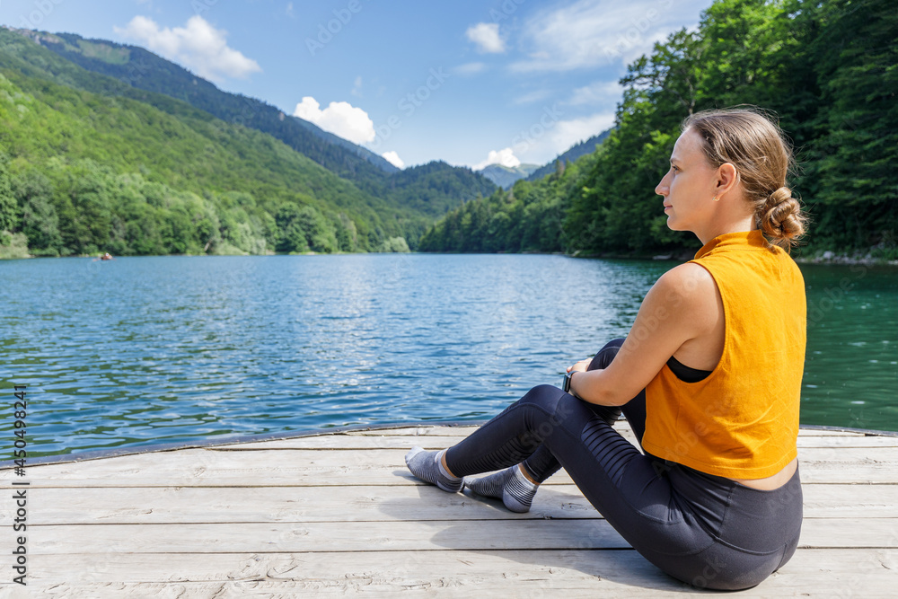 Young sporty fitness woman sitting on the pier near the mountain lake.