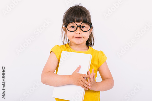 Smiling little girl wearing yellow t-shirt and round black glasses holding a book in her hands on white background