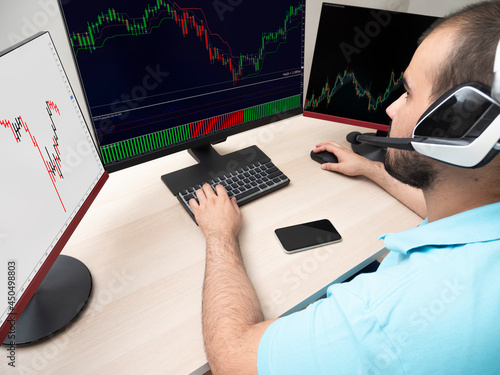 Young caucasian man in a blue shirt sitting with a table and a computer with 3 monitors doing trading with headphones on your head