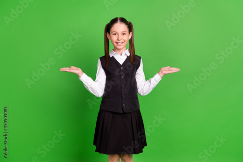 Photo of pretty funny school girl wear black white uniform compare arms empty space smiling isolated green color background