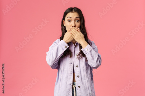 Portrait of surprised young girl found out something she shouldnt have, cover mouth as gasping and staring at camera speechless, trying not slip secret, gossiping, pink background photo