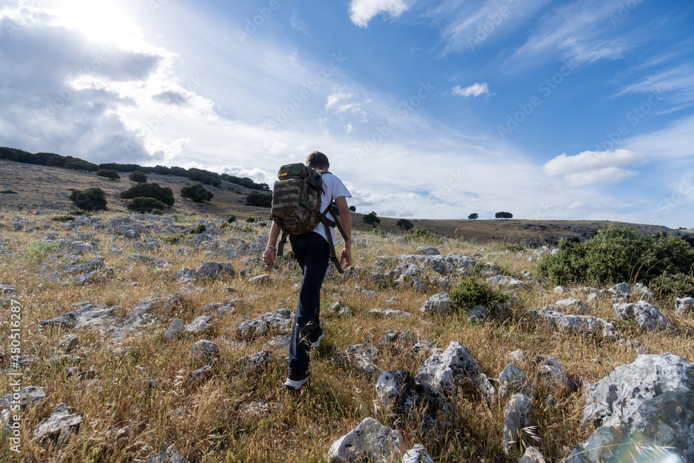 Rear side view of a teenager with big backpack hiking through rocky valleys on a sunny day with a cloudy sky