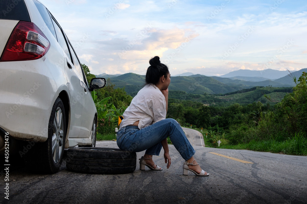 Young Asea woman sitting near the car for calling for help on the public road in the forest area at mountain and sky background