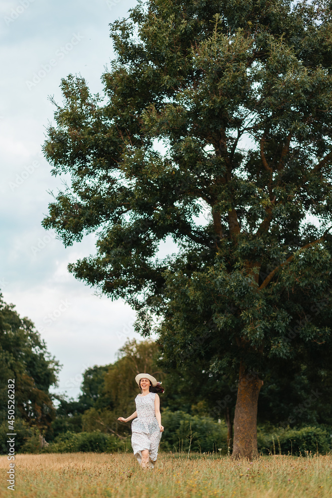 Summer holidays. A happy young woman in a straw hat and a white dress runs across the field. Vertical