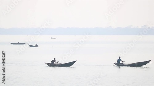 Men sitting boats paddle along the Padma river in hazy sunshine.