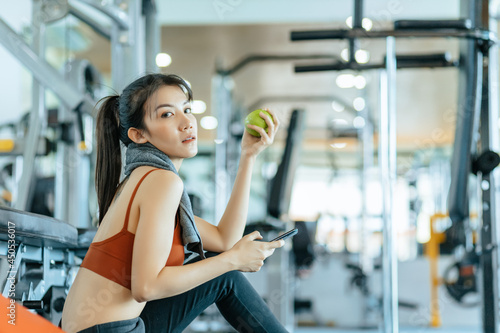 Woman exercise in gym fitness breaking and relax. hand  holding apple fruit after training sport and dumbbell, water bottle on the ground.
