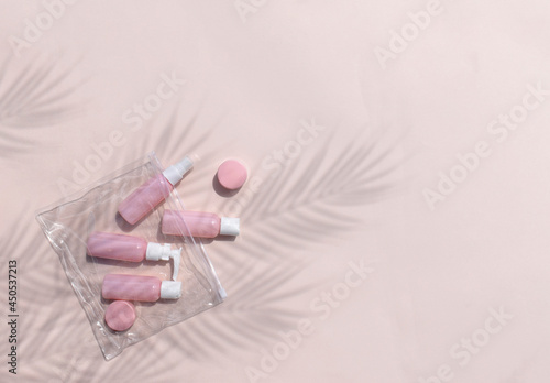 Mock up feminine flatlay with small size cosmetic bottles in travelling bag on neutral pastel background. Palm leaves shadows. Minimal skin or body care products concept for vacation or journey.