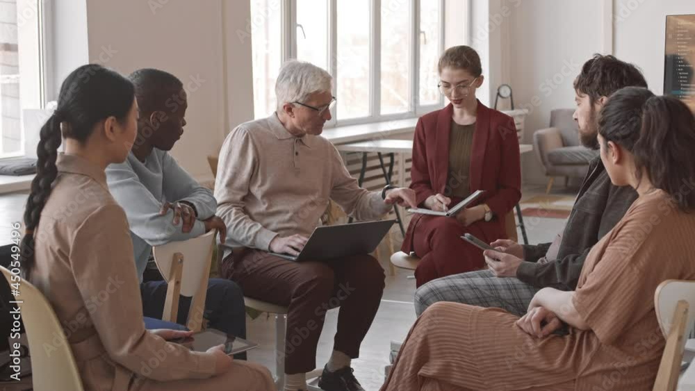 Medium long of multiethnic female and male marketing specialists sitting in circle in office, talking, having business meeting Medium long of multiethnic female and male marketing specialists sitting in circle in office, talking, having business meeting