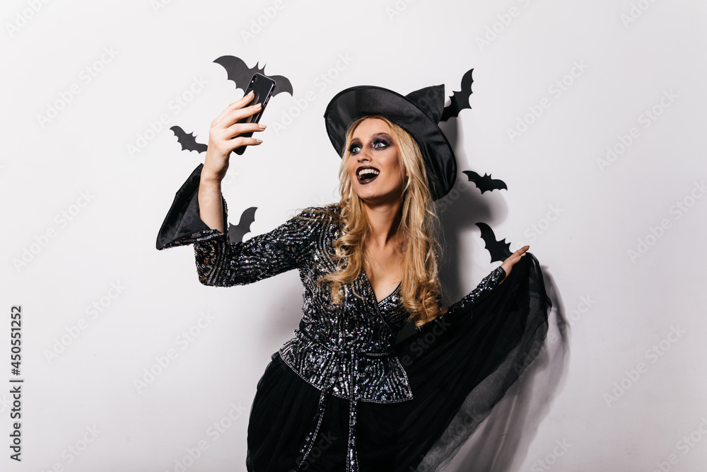 Laughing carefree girl making selfie with halloween bats. Studio portrait of pleasant young witch dancing at party. Laughing carefree girl making selfie with halloween bats. Studio portrait of pleasant young witch dancing at party.