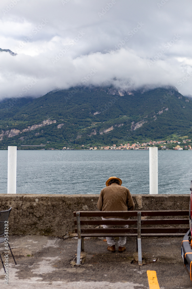 An Italian man with a hat sitting on a bench by Lake Como
