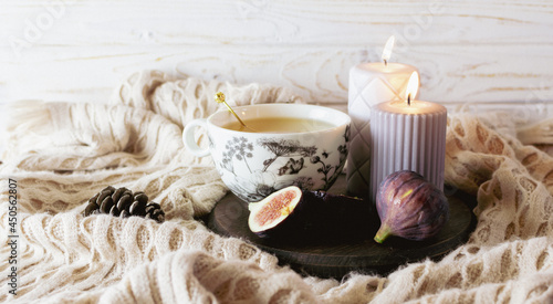  Creative hygge style composition with cup of herbal tea, sweet ripe figs, candles and warm plaid on white wooden background. Cozy home interior.